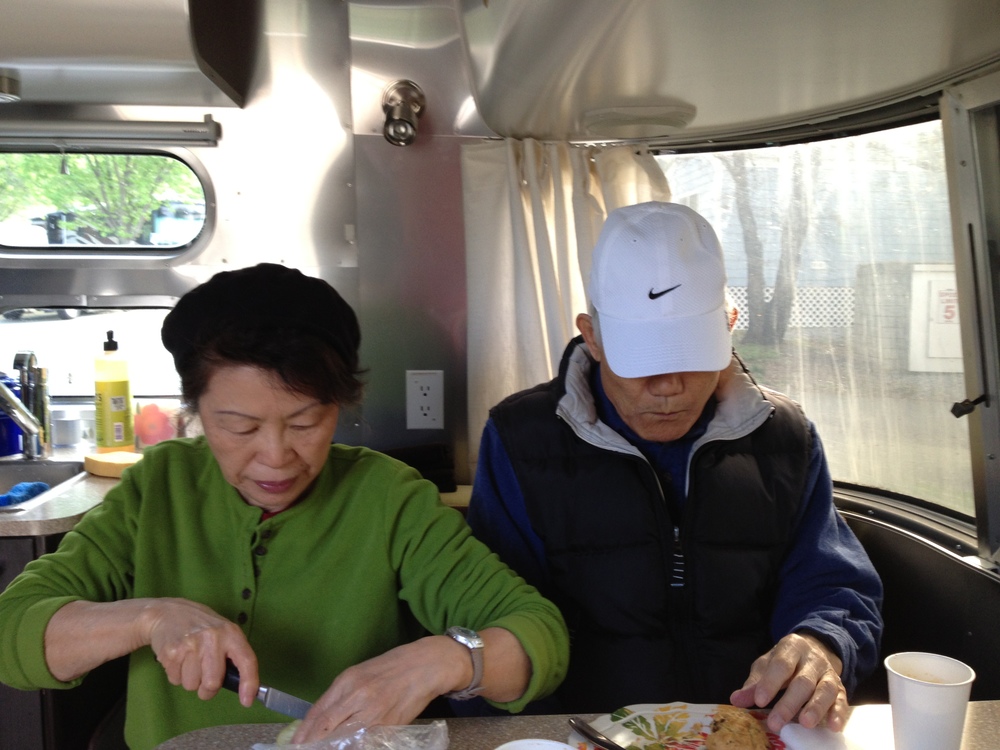 Two individuals sitting inside a trailer, one preparing food and the other eating, with a window revealing a natural view.