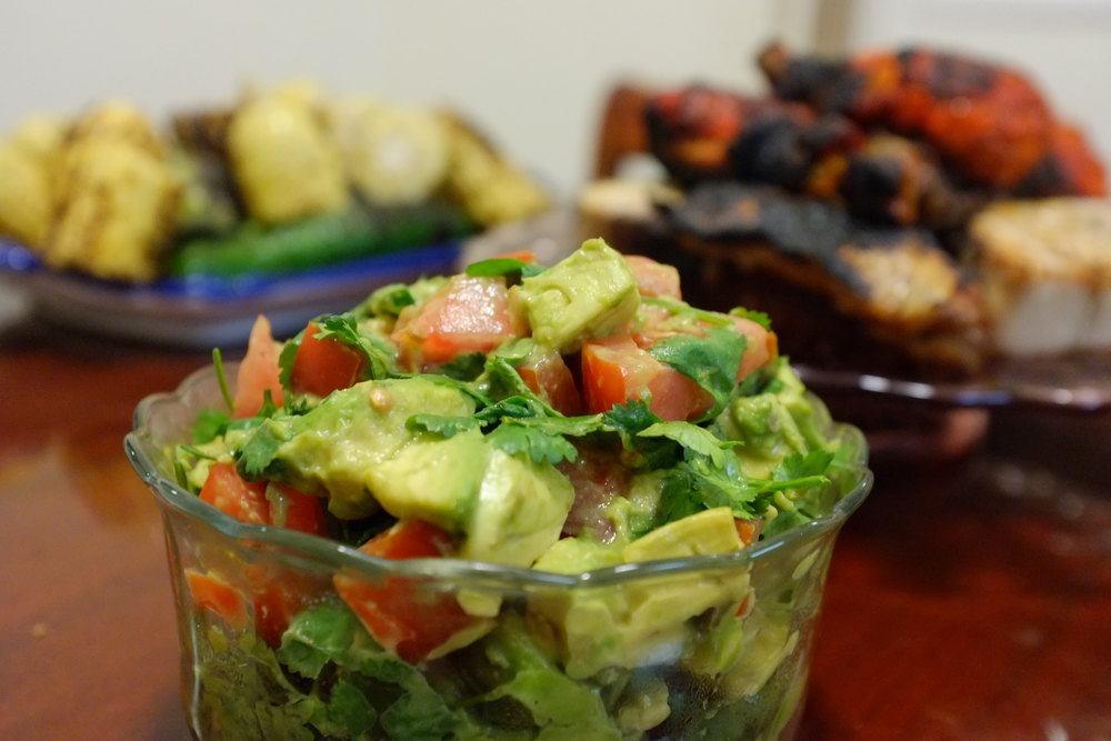 A bowl of fresh guacamole made with avocado, tomatoes, and cilantro, placed in the foreground with grilled food in the background.