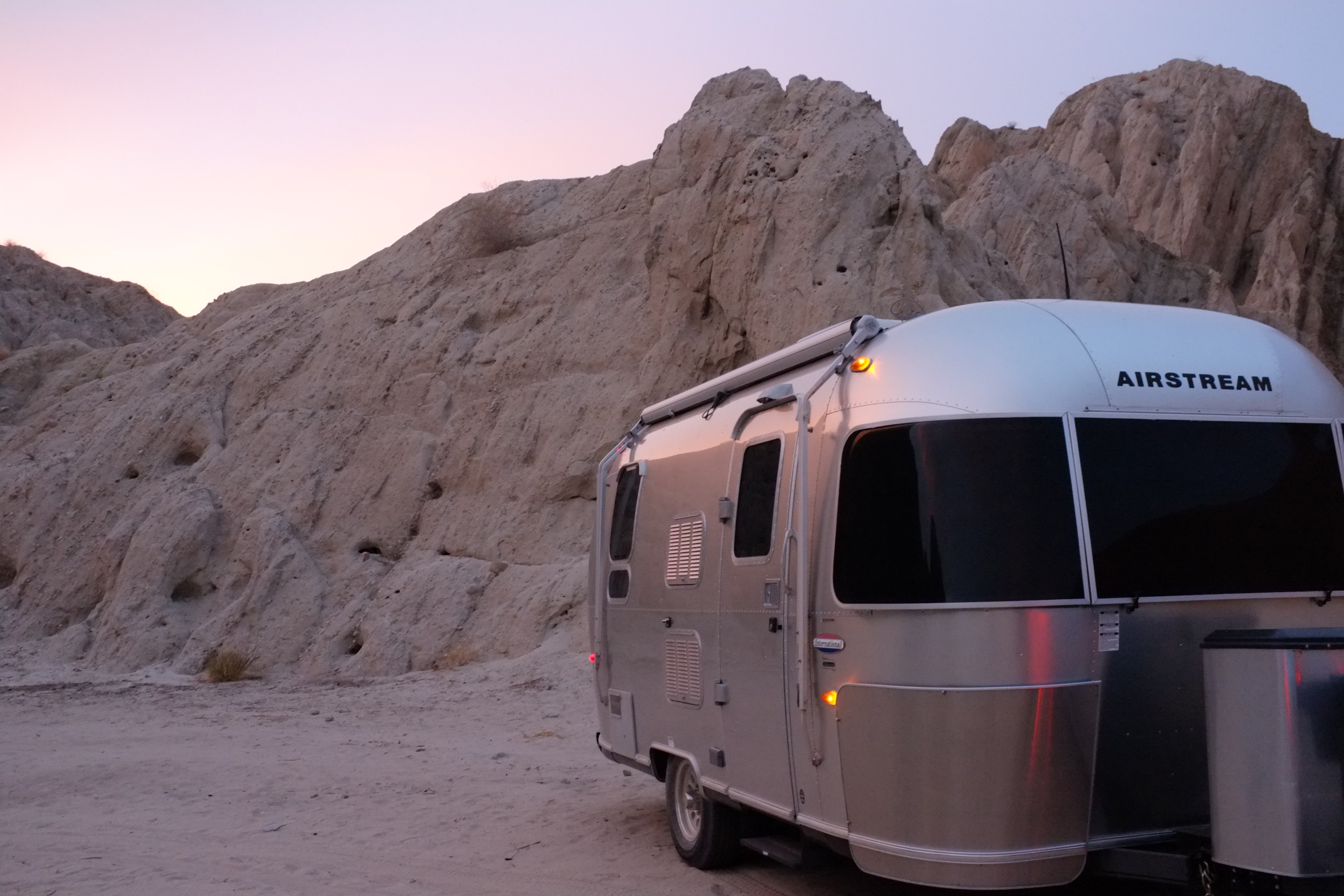 A silver Airstream trailer parked in a sandy, rocky landscape at dusk, with pink and purple hues in the sky.