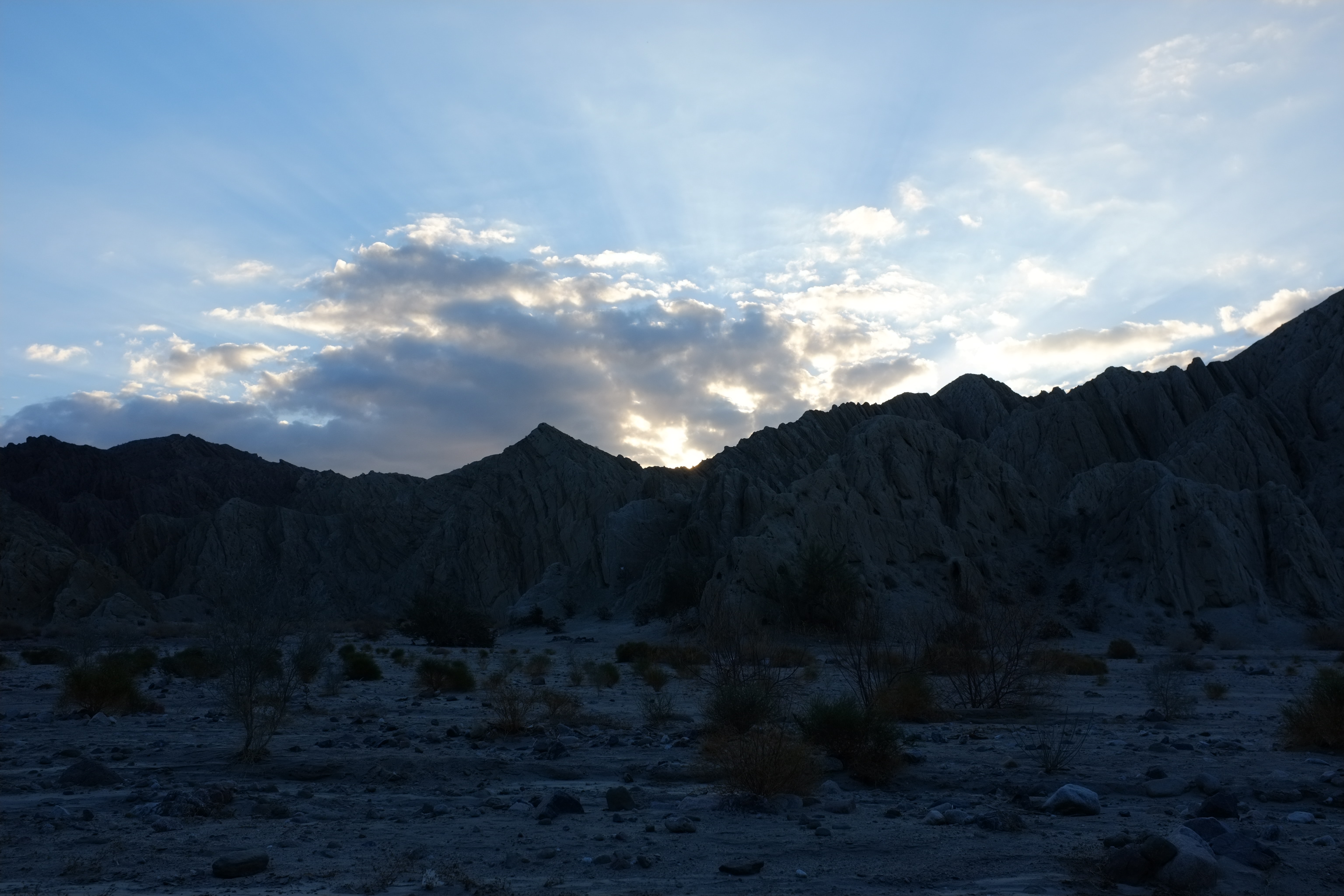 A dramatic landscape photograph featuring silhouetted mountains against a cloudy sky at sunset, with rays of light emerging from behind the peaks.