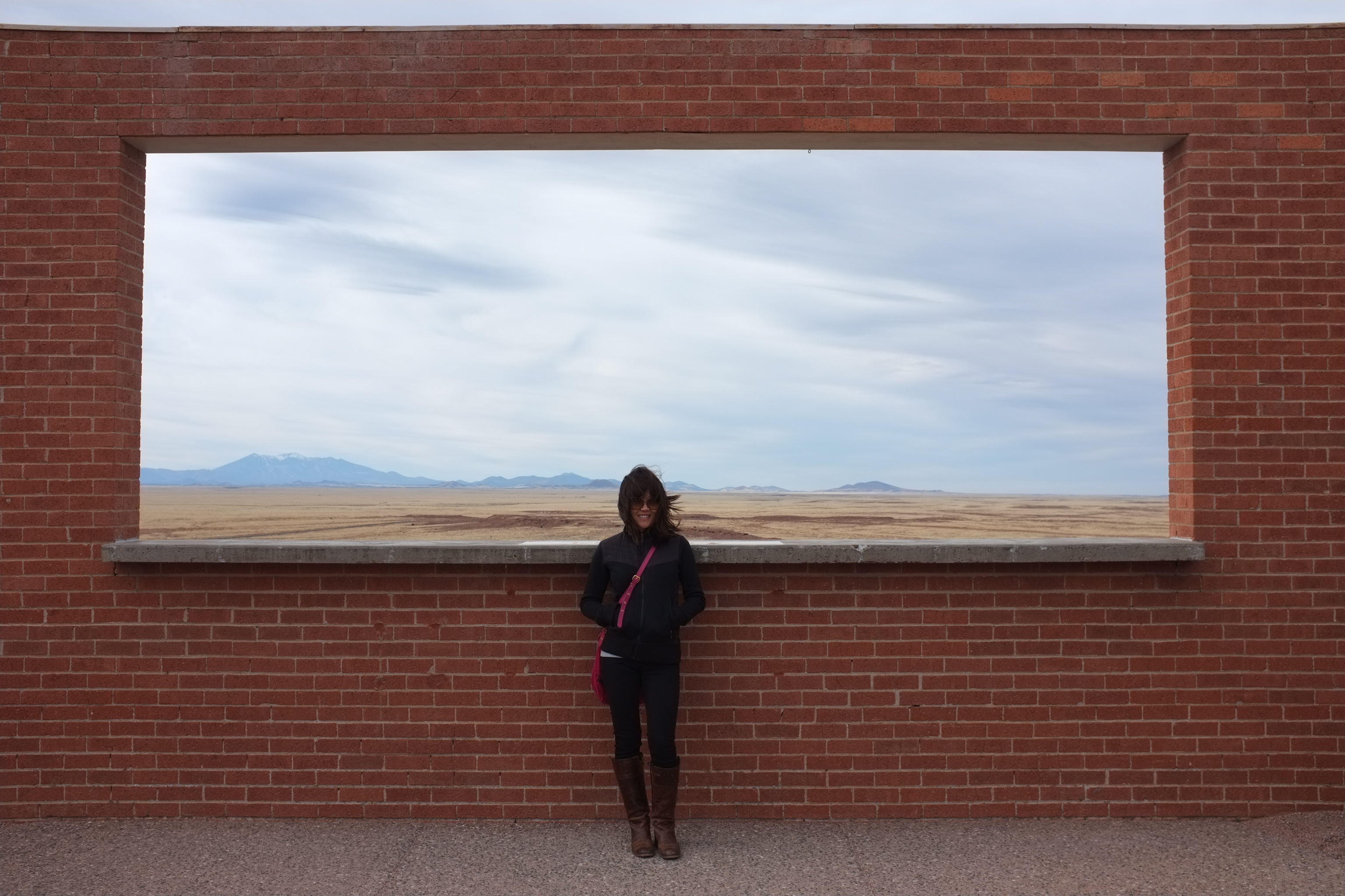 A woman stands in front of a brick wall with a rectangular opening, overlooking a vast landscape with mountains in the distance and a cloudy sky.