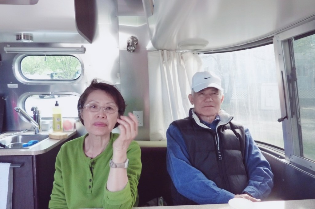 A woman in a green top and glasses sits at a dinette table inside a trailer, gesturing with her hand. An older man in a blue shirt and white cap sits beside her, looking serious. The interior features windows and a kitchenette area.
